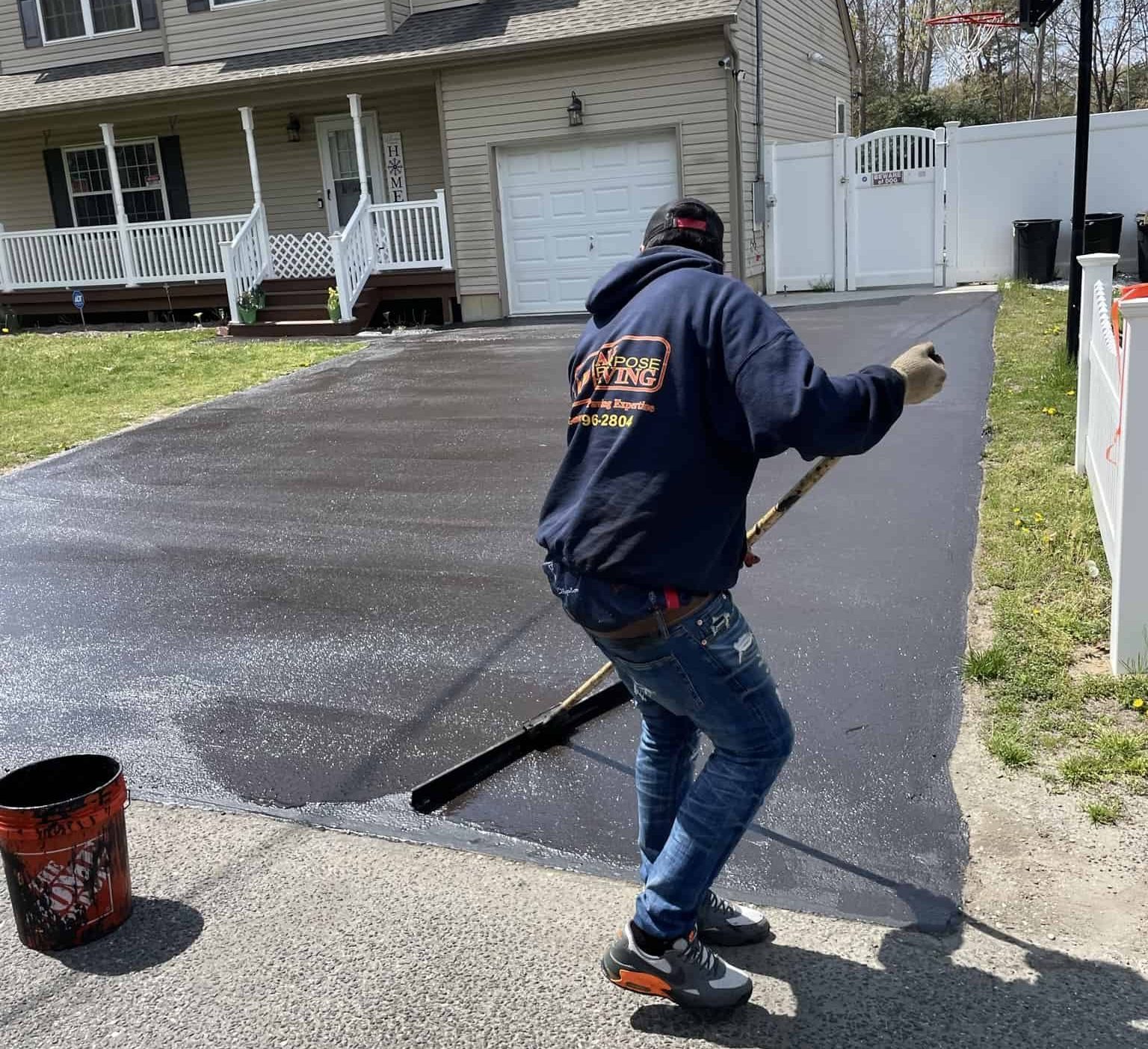 A person in a hoodie and jeans spreads black sealant on a driveway with a long-handled tool, applying Driveway Coating in Suffolk County, NY, in front of a suburban house with a porch and garage on a sunny day.
