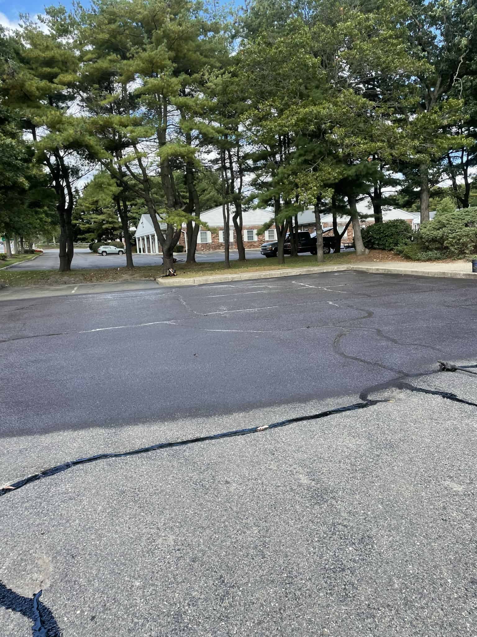 A mostly empty parking lot with a few parked cars near the edge, bordered by trees and shrubs. In the background, a white building is partly visible through the trees under a partly cloudy NY sky—ideal for commercial paving in Suffolk County.