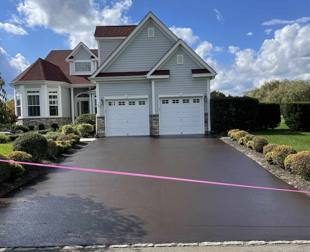 A two-story suburban house with a fresh black driveway coating in Suffolk County, NY, bordered by shrubs and a pink caution tape across the entrance, under a blue sky with white clouds.