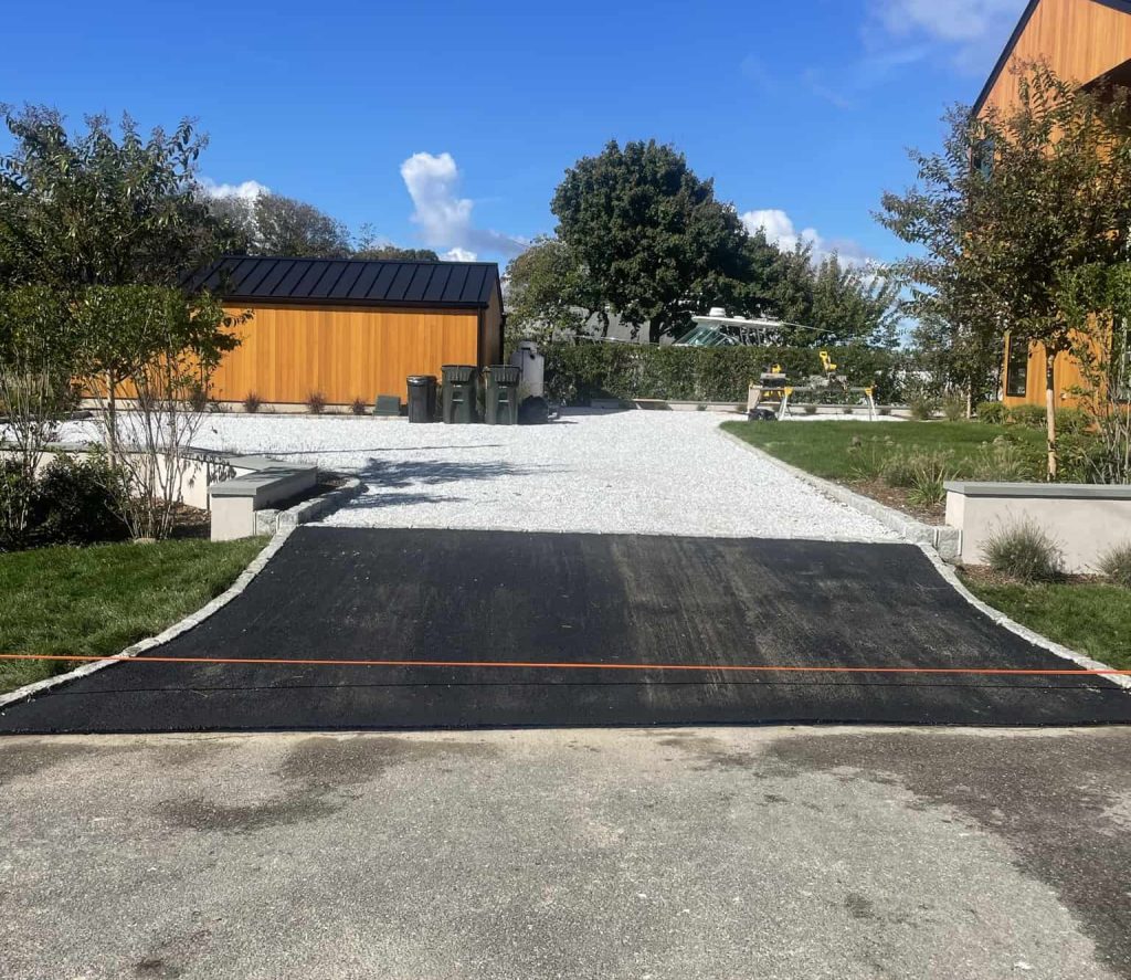 A driveway with a dark asphalt ramp, crafted by Residential Paving in Suffolk County, leads to a gravel parking area between two modern orange buildings in NY, surrounded by green grass, trees, and a bright blue sky.
