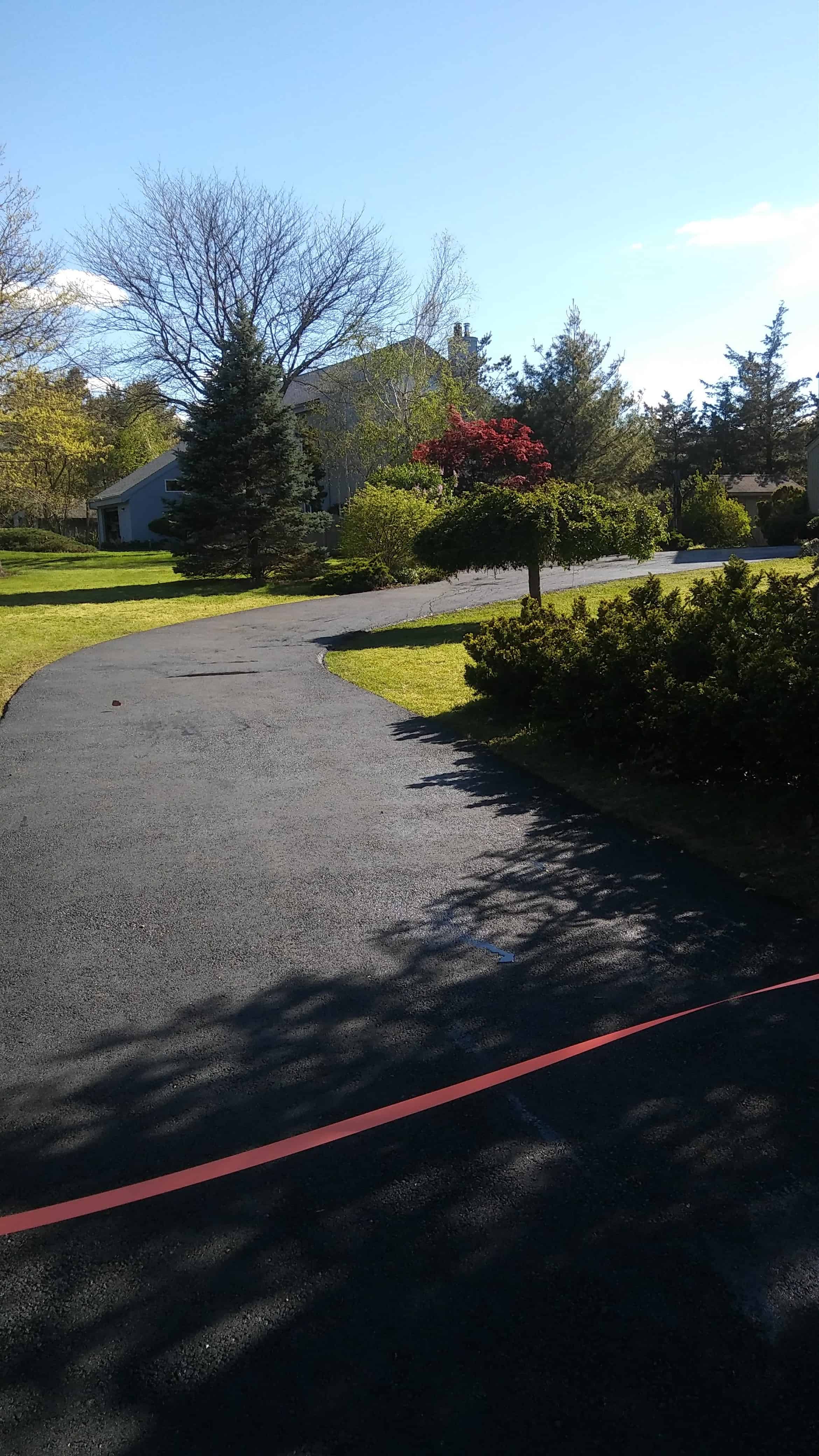 A paved driveway curves through a green yard with trees and shrubs, leading to a house partially visible in the background. A red line marks the freshly applied driveway coating in Suffolk County, NY, under a clear blue sky.
