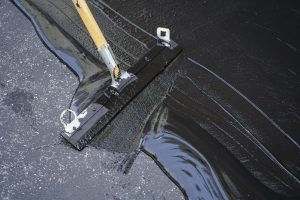 A close-up of a broom with a wooden handle spreading black sealant over cracked asphalt, smoothing the surface during a Seal Coating in Suffolk County, NY pavement repair or maintenance process.