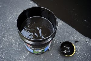 An open black bucket filled with thick black liquid, possibly asphalt or tar, sits on a gray textured surface. The bucket's lid is off and placed nearby, resting upside down—typical for Asphalt Services in Suffolk County, NY.