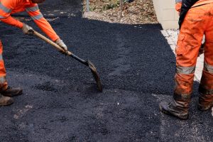 Two workers in orange reflective clothing spread fresh asphalt with a shovel, resurfacing a roadway or driveway. Their legs and lower bodies are visible, and the ground is covered with dark, newly laid asphalt.