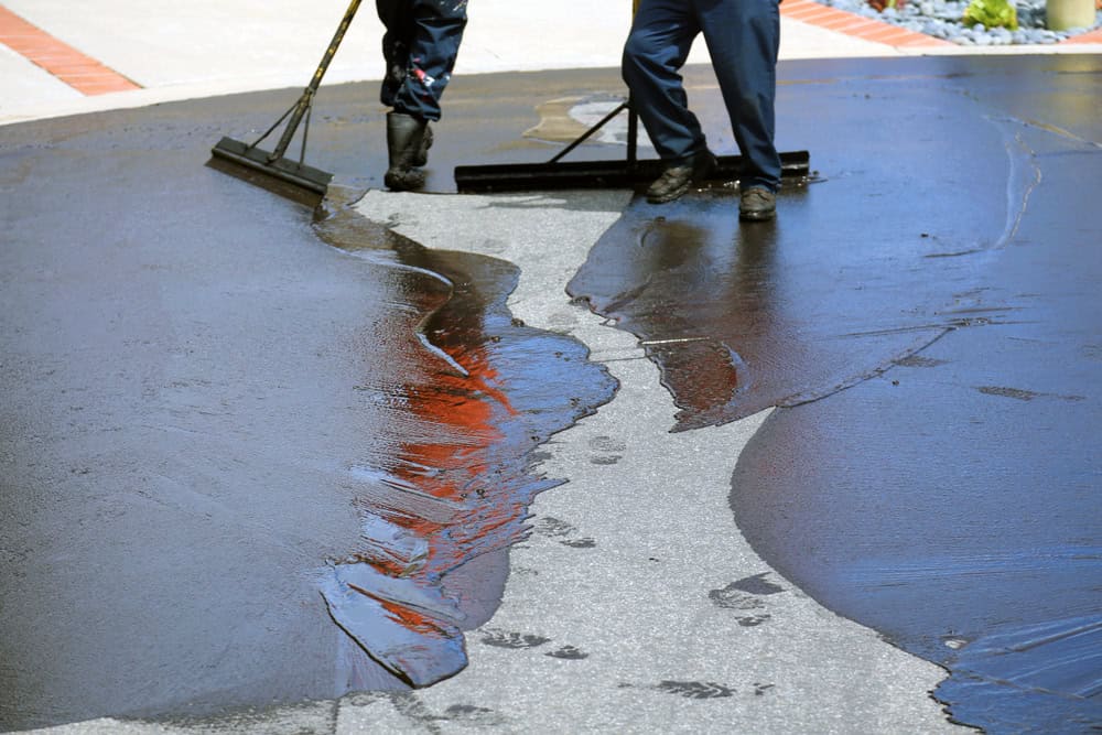Two people spreading liquid sealant on a driveway with large brushes, ensuring an even coat to protect the surface.