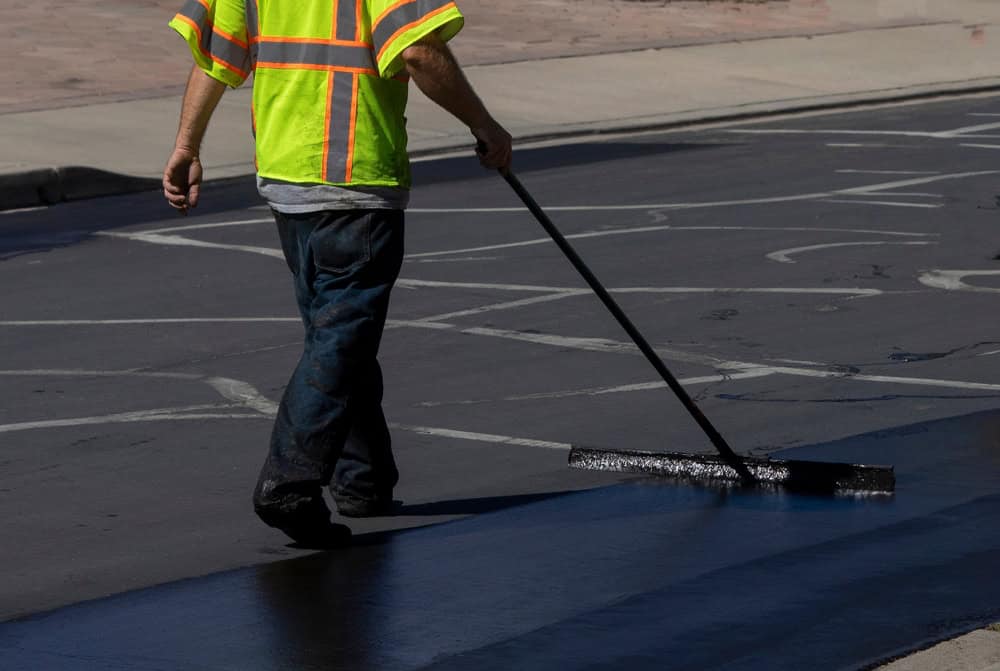 A worker in a neon yellow safety vest uses a long-handled tool to spread asphalt on a Suffolk County, NY road. The surface is dark and smooth, indicating recent treatment, suggesting ongoing asphalt paving maintenance work.
