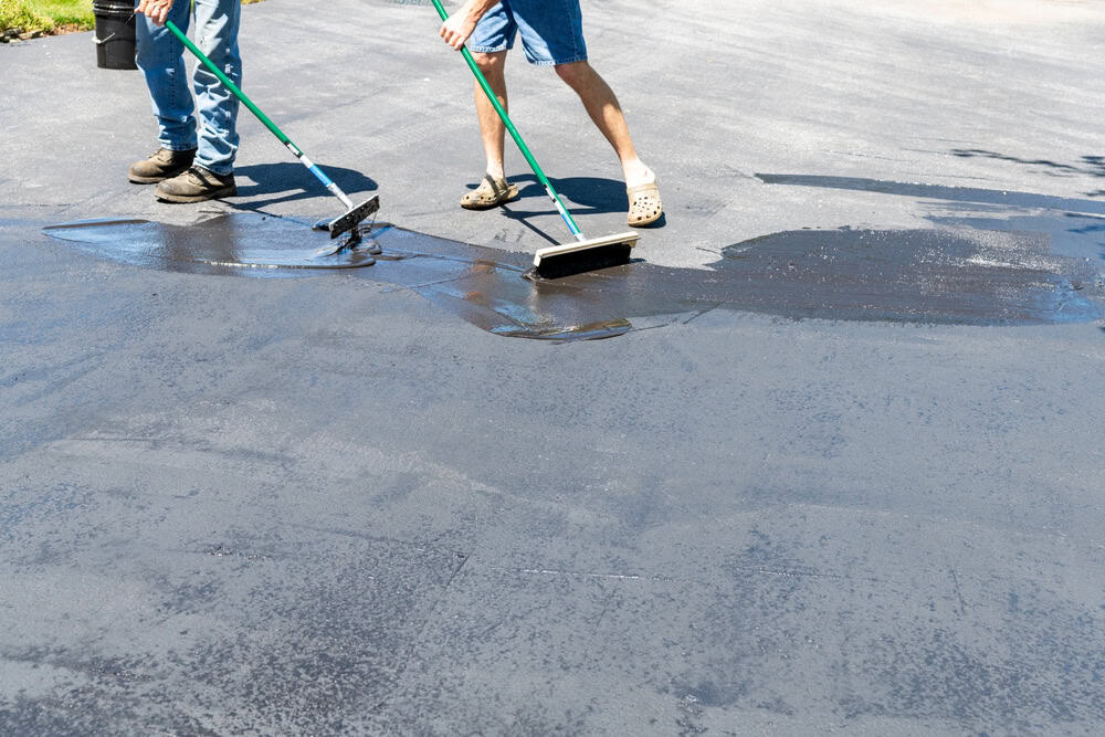 On a sunny day, two people in casual clothes are applying sealant to a driveway with long-handled brushes. Their meticulous work brings to mind the skilled touch of professionals specializing in asphalt paving in Suffolk County, NY.