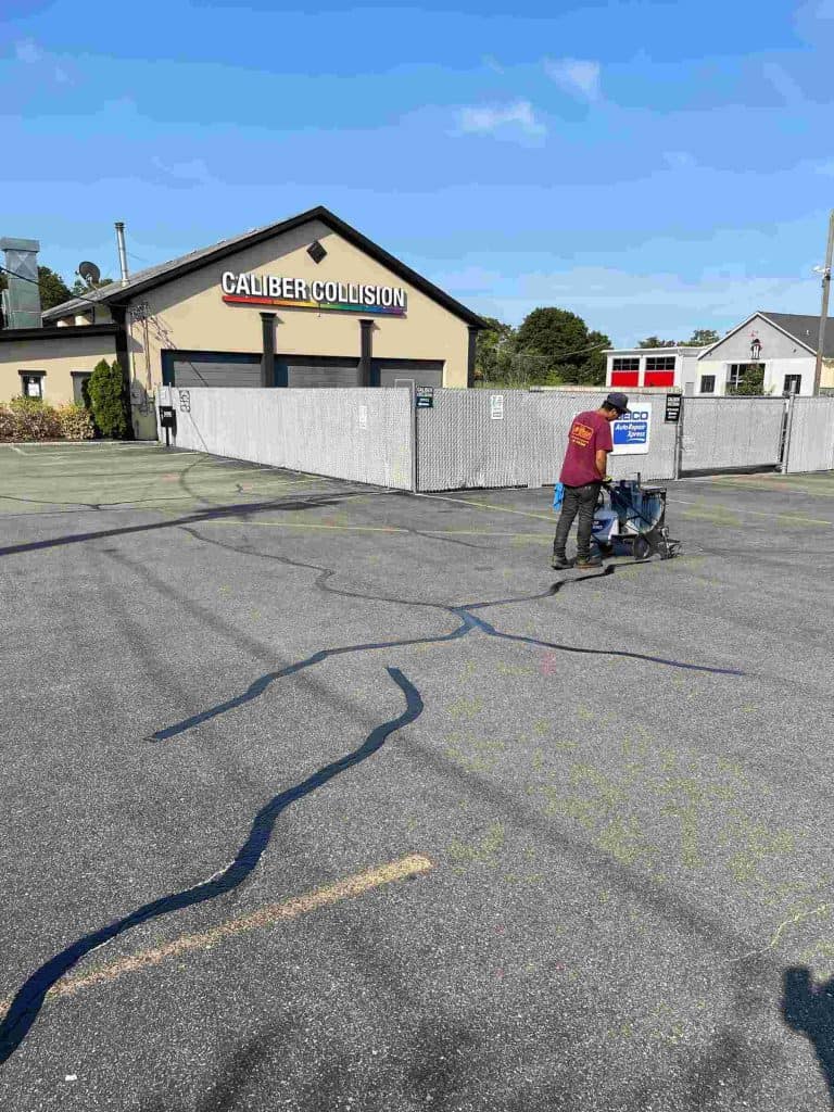 A person pushes a shopping cart through an empty parking lot in front of a building labeled "Caliber Collision." The scene is under a clear blue sky, with a few other buildings and trees in the background. Long shadows stretch across the ground.