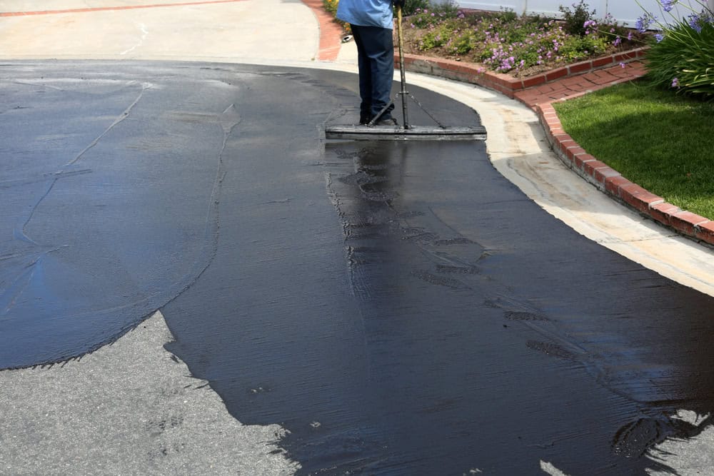 A worker spreads sealant on a driveway with a large applicator. The wet, black sealant contrasts with the dry, lighter-colored concrete. The driveway is bordered by a brick edge and flowering plants.