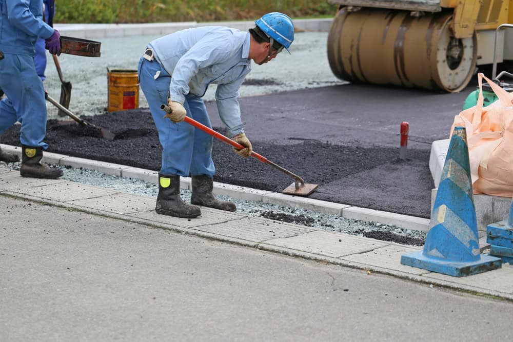 Worker in blue uniform and helmet smooths asphalt with a shovel on a construction site. A roller machine is in the background, and traffic cones and construction materials are nearby.