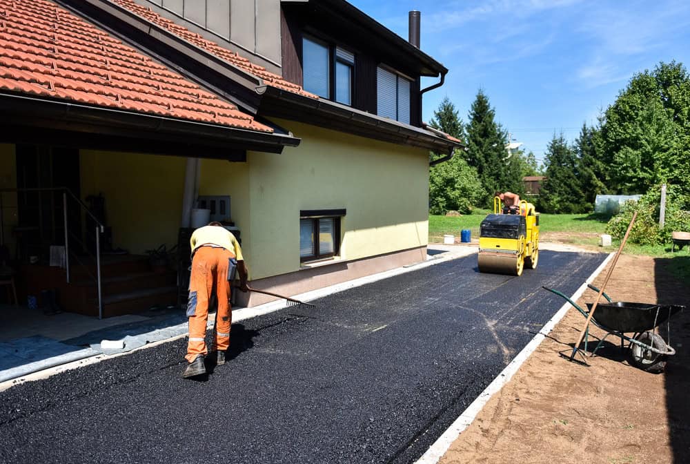 Worker in orange clothing is smoothing newly laid asphalt on a driveway next to a house. A small yellow steamroller is parked on the freshly paved surface. A wheelbarrow and tools are nearby. Sunny day with green trees in the background.