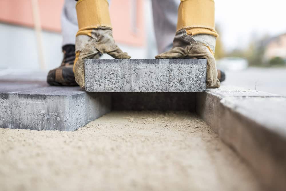 A person wearing work gloves lays a concrete block on a bed of sand, aligning it with a similar block already placed. The person appears to be paving a surface, with a building and a blurred background visible.