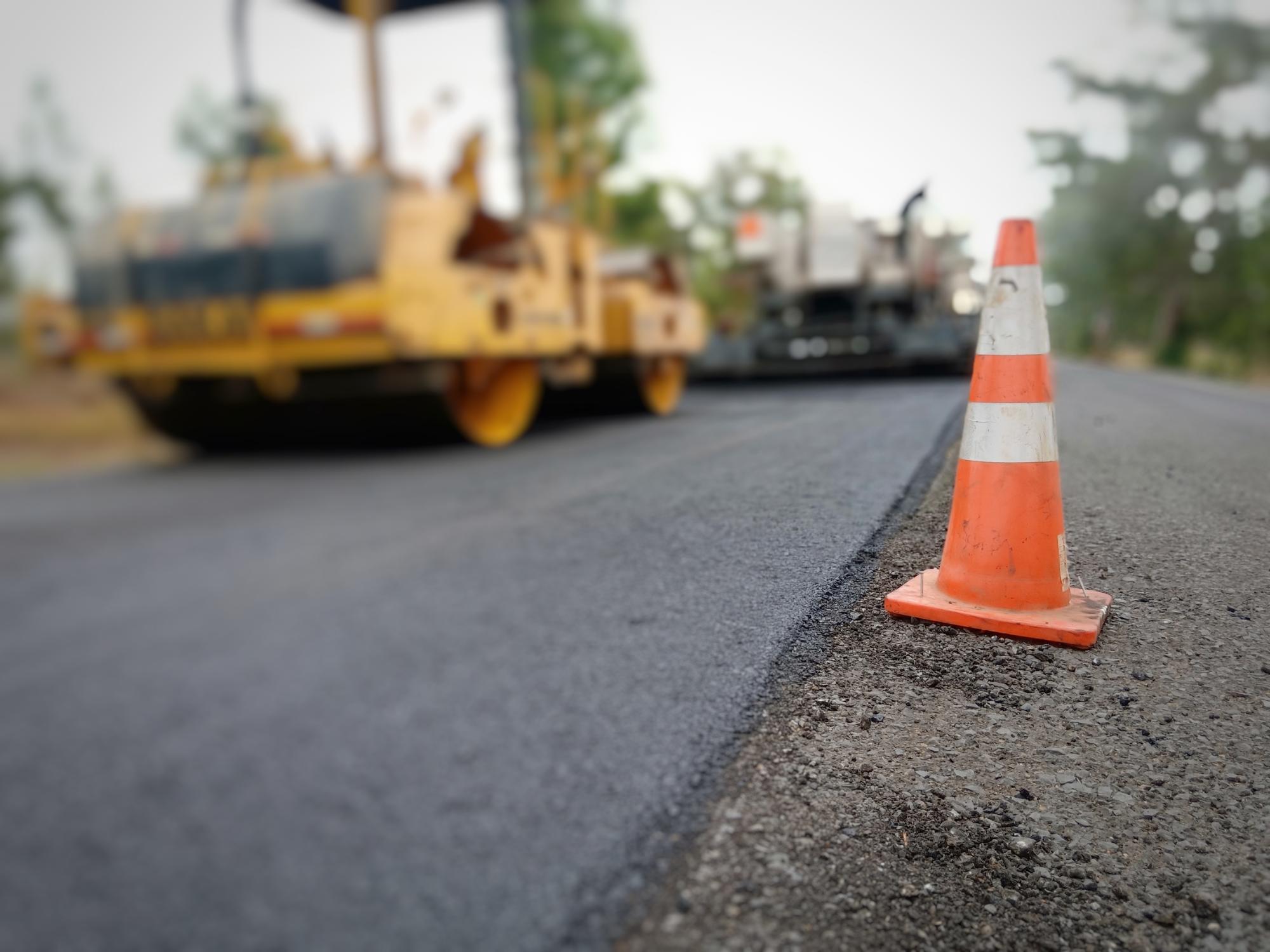 An orange traffic cone is placed on the edge of a freshly paved road, with a blurred construction vehicle in the background.