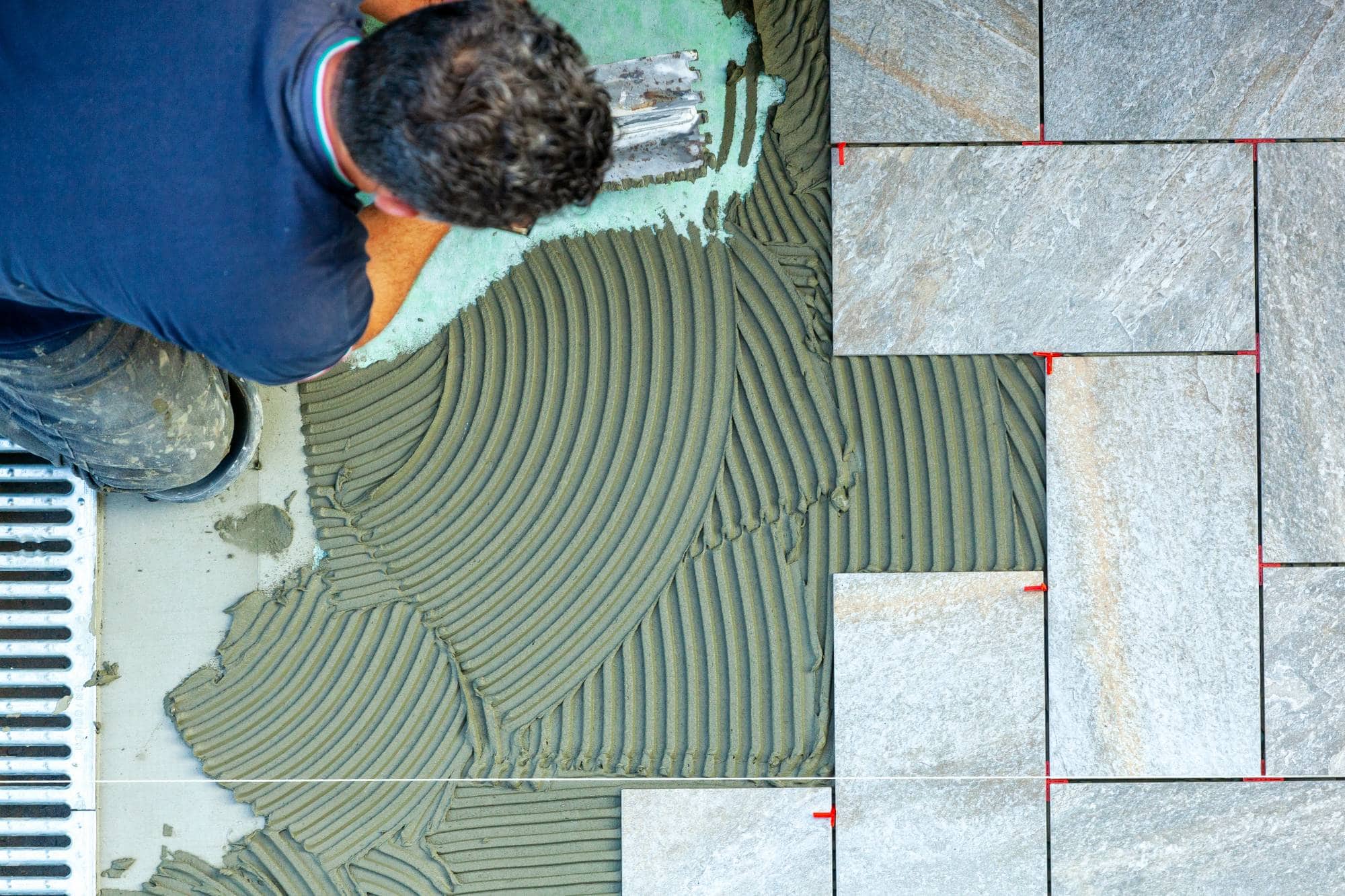 A person is applying grey tile adhesive with a notched trowel, laying large rectangular tiles in a staggered pattern. Red spacers are visible between the tiles.