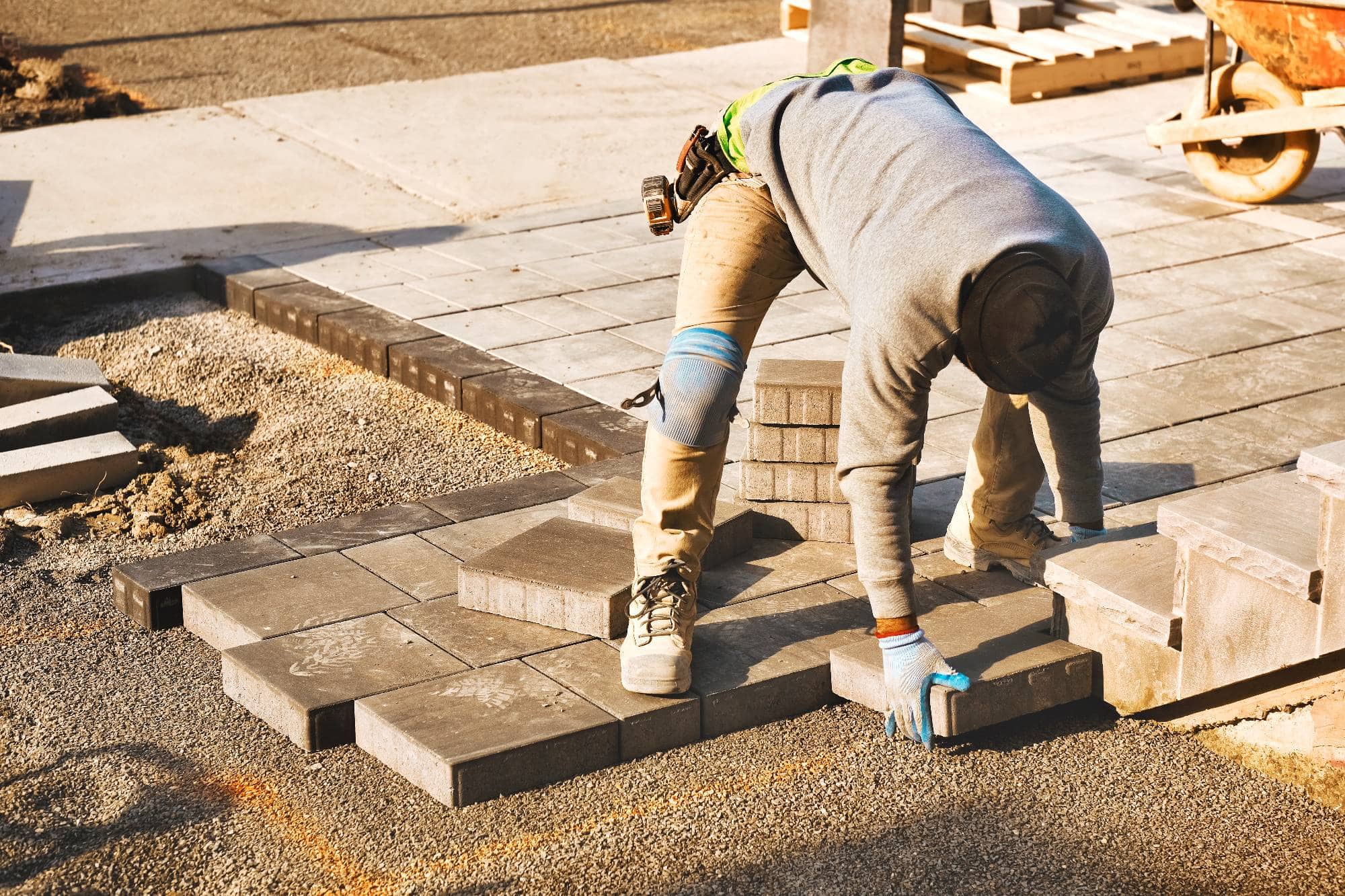 A construction worker in a gray hoodie and gloves is bending over, placing rectangular paving stones on the ground to build a sidewalk or patio. Tools and construction materials are visible in the background.