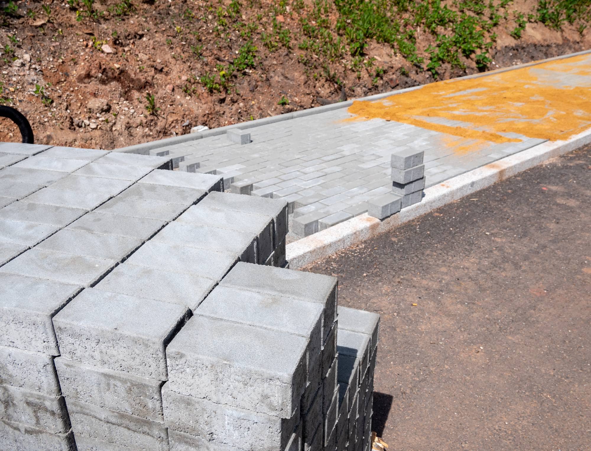 Stacks of gray concrete paving stones on the ground next to an unfinished pathway, with some pavers already laid in place; yellow sand and soil are visible nearby.