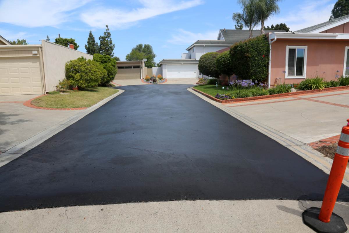 A freshly paved, black asphalt driveway leads up between two suburban houses with manicured lawns and garages; a bright orange traffic cone stands at the edge of the new pavement.