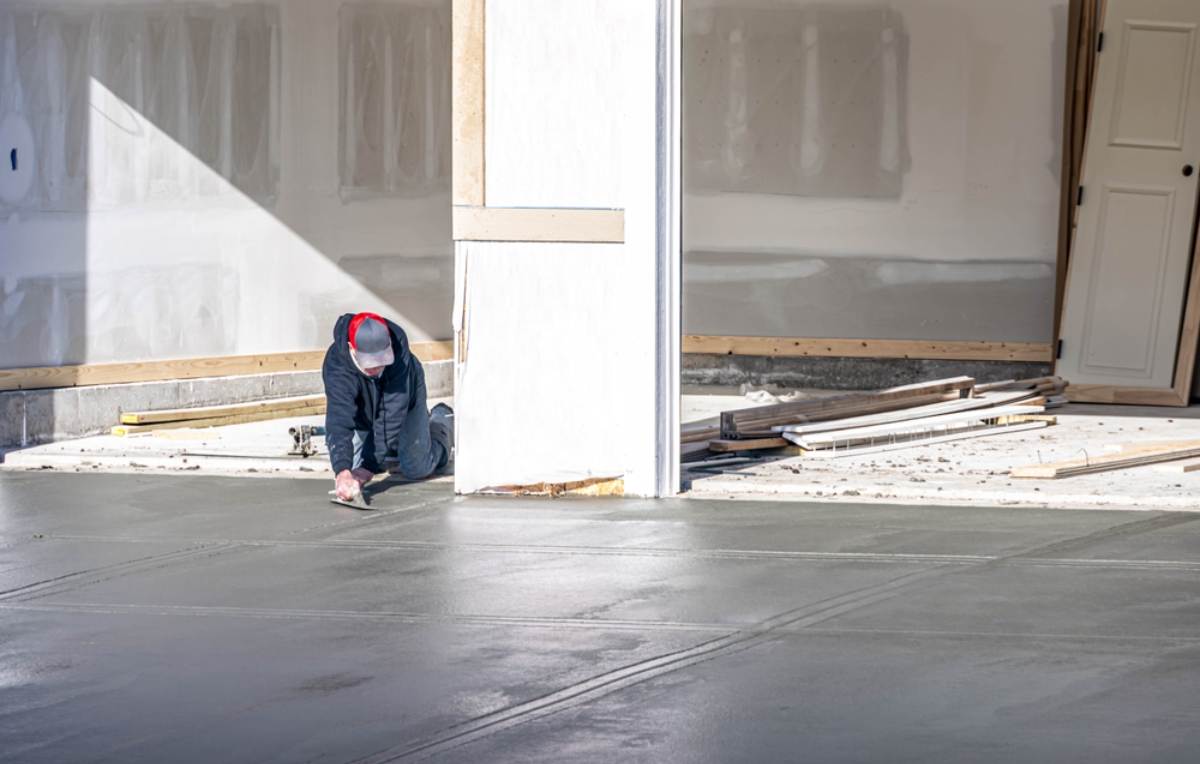 A worker wearing a hoodie and mask smooths wet concrete with a trowel near a garage under construction; drywall and building materials are visible in the background.