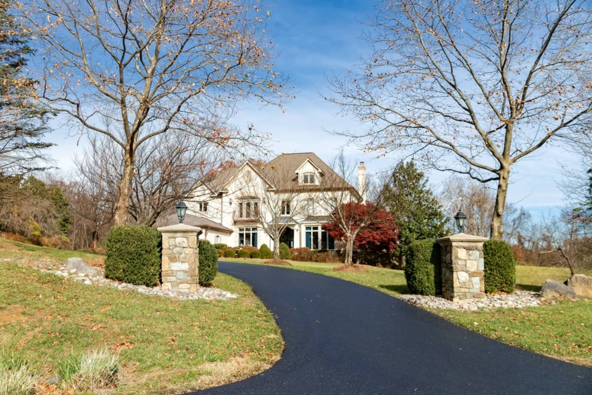 A large suburban house with a peaked roof sits at the end of a curved, freshly paved driveway, surrounded by trees and landscaped bushes under a clear blue sky.