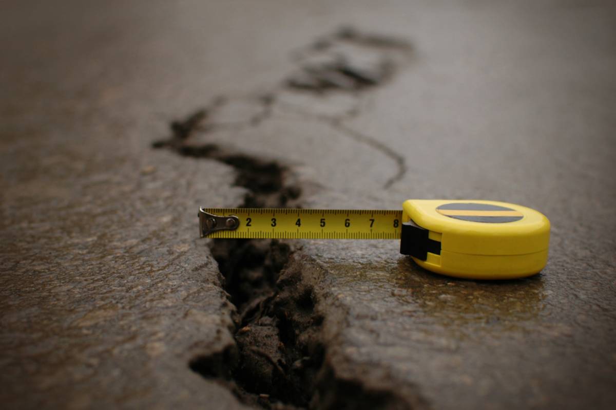 A yellow tape measure is extended across a crack in an asphalt surface, showing the width of the crack. The image focuses on the crack and the measuring tape.