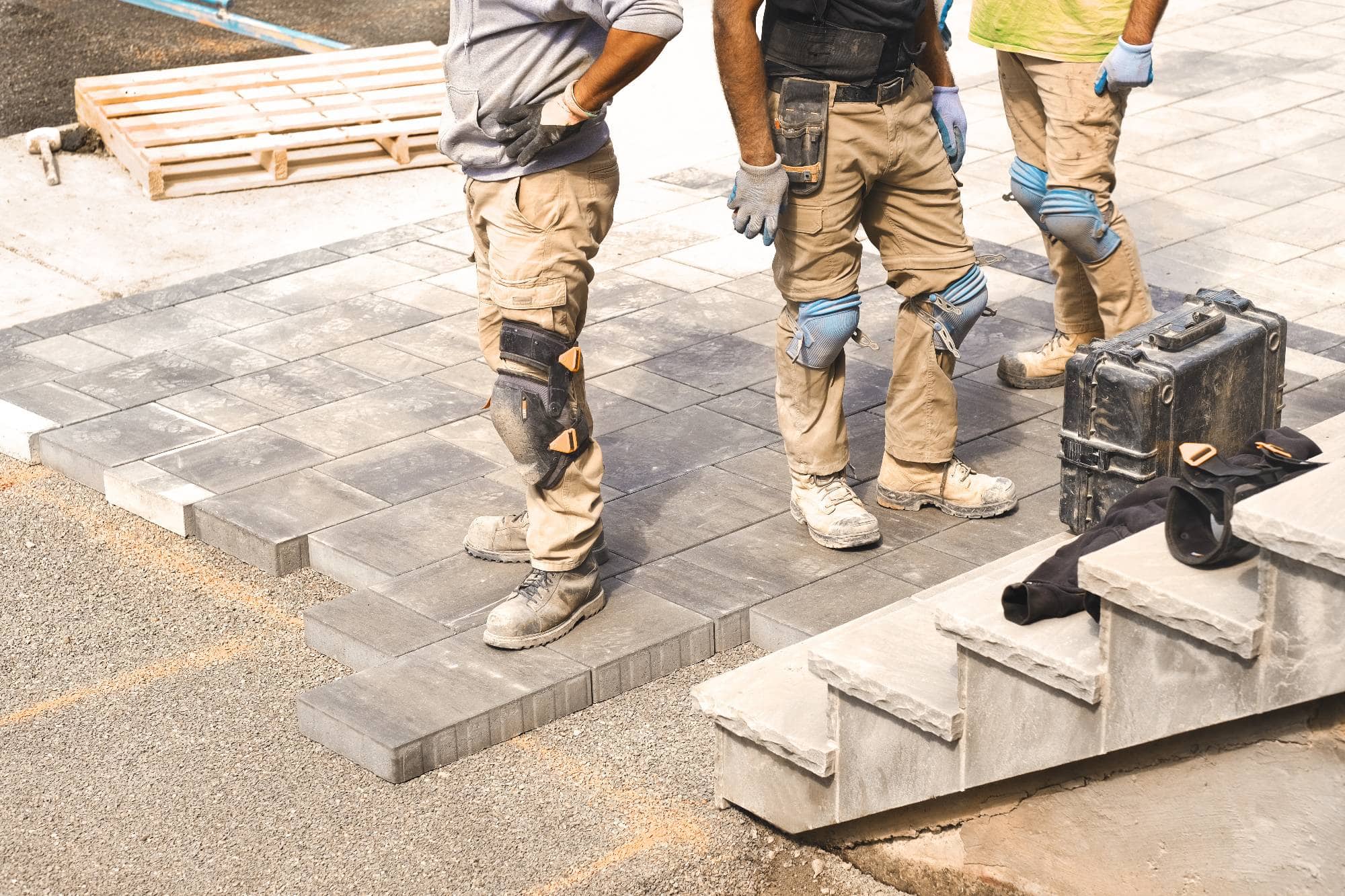 Three construction workers in work clothes and kneepads stand on a partially completed outdoor stone patio near steps, with tools and a black toolbox nearby. Their upper bodies are mostly out of frame.