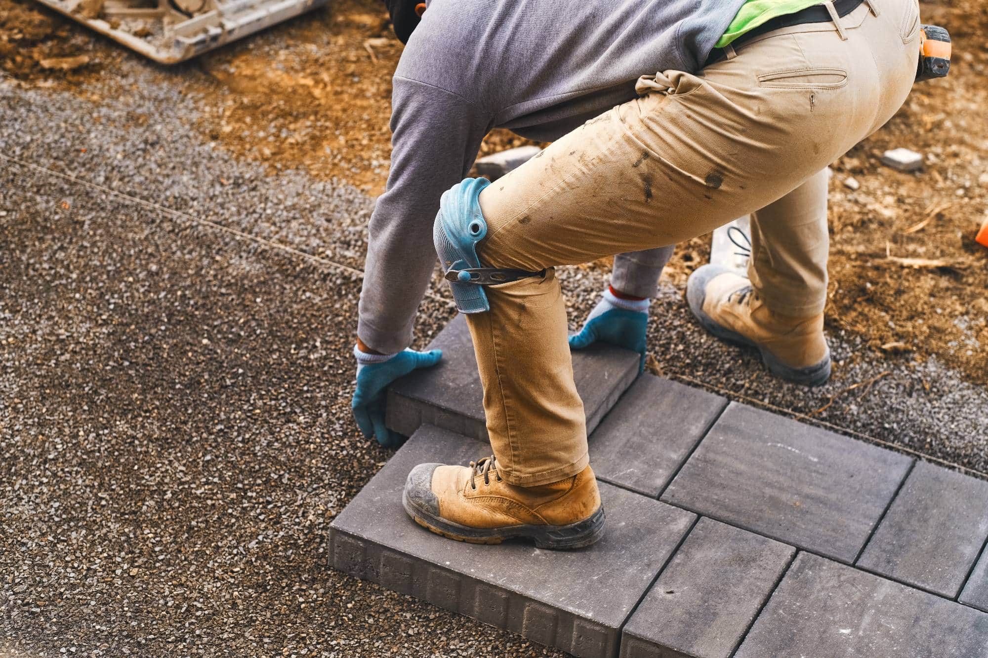 A construction worker wearing gloves, knee pads, and boots is crouched down, placing gray paving stones onto gravel at a work site. Dirt and tools are visible in the background.