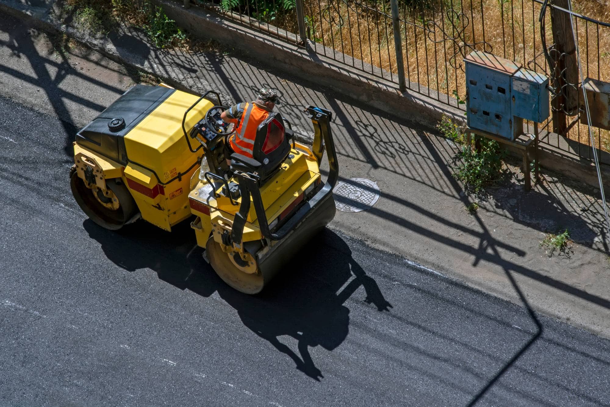 A construction worker wearing an orange safety vest operates a yellow road roller on freshly paved asphalt near a metal fence and utility boxes, casting a clear shadow on the ground.
