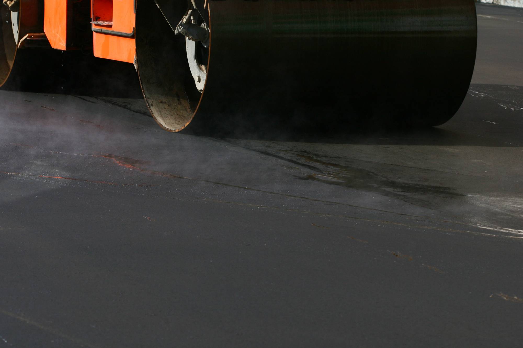 A close-up of a steamroller compacting fresh, steaming asphalt on a road surface. The lower part of the orange machine and its large metal drum are visible.