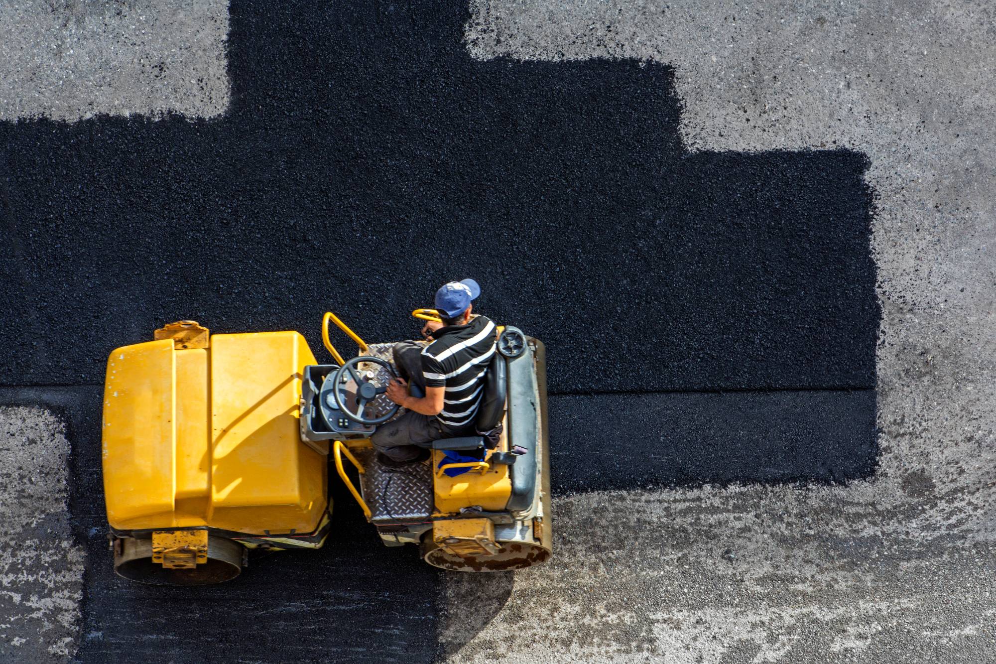 A person in a striped shirt operates a yellow road roller, compacting black asphalt onto a gray road surface, as viewed from above.