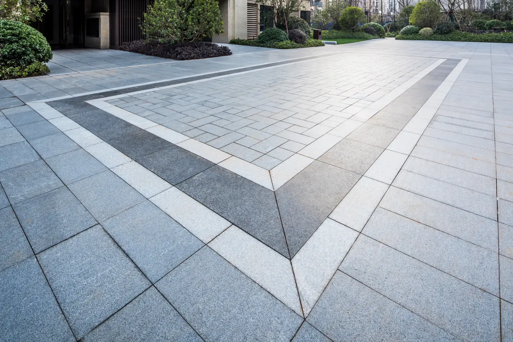 A modern, spacious outdoor area with grey stone tiles arranged in a geometric pattern, featuring a large triangle outlined by darker and lighter tiles, surrounded by neatly trimmed bushes and greenery.