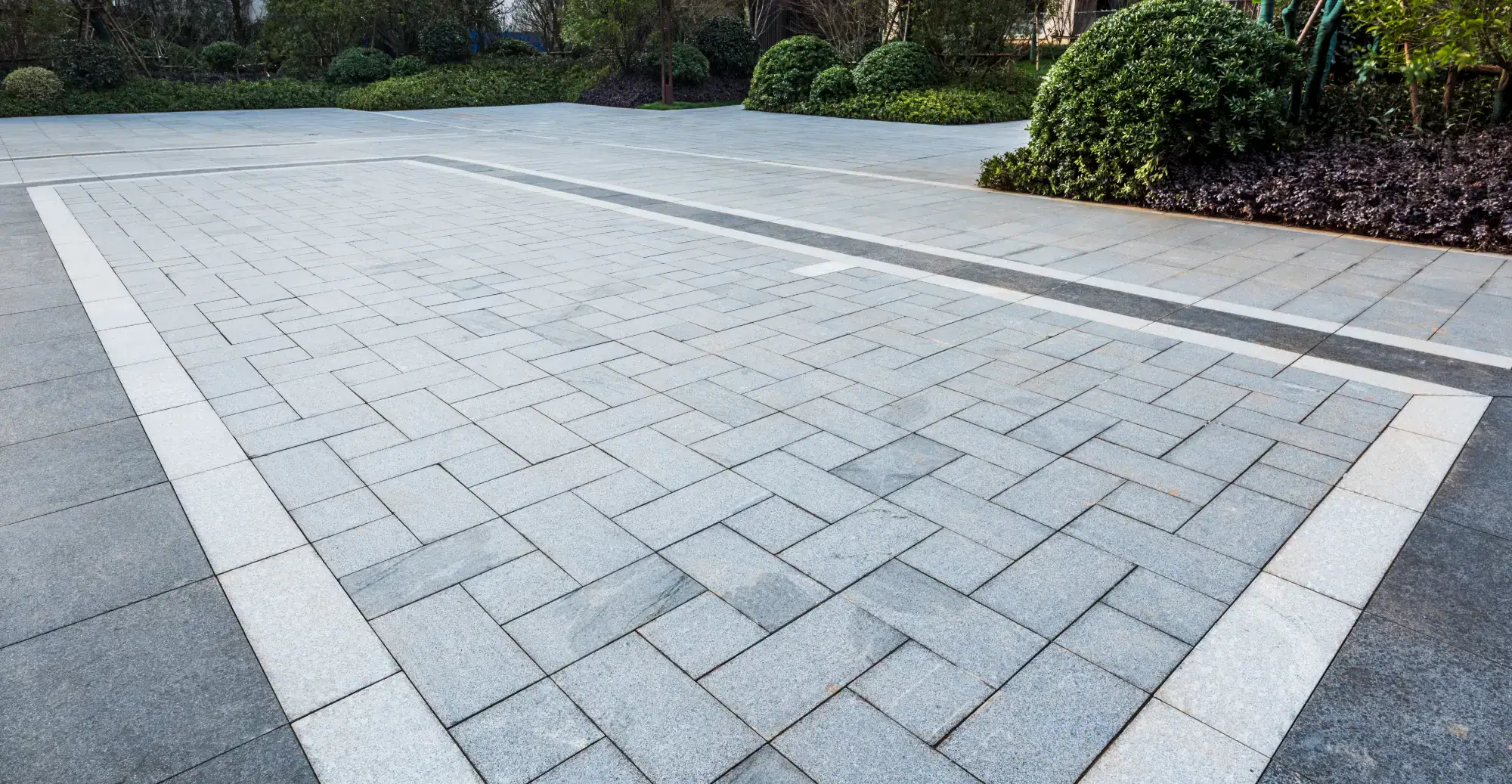A spacious outdoor area with neatly arranged gray paving stones, bordered by greenery, bushes, and trees in the background.